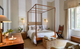 Elegant bedroom featuring a wooden four-poster bed, soft bedding, and neutral decor. The room includes a wooden dresser, a cozy chair, and natural light streaming through large windows, creating a serene atmosphere.