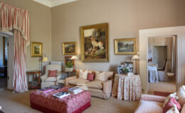 Elegant living room featuring plush seating, decorative pillows, and an ornate coffee table. The walls are adorned with classic paintings, and large windows are dressed with flowing drapes, creating a warm and inviting atmosphere.