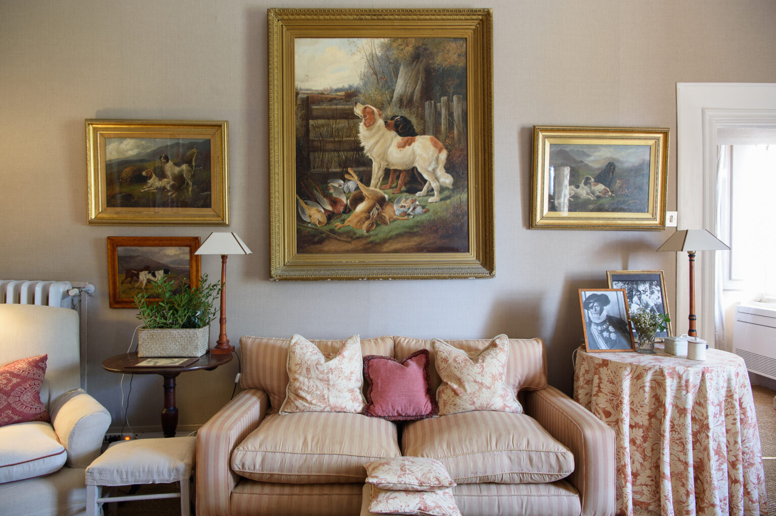 Cozy living room featuring a vintage sofa with decorative pillows, surrounded by framed animal paintings on the walls. A side table with a lamp, potted plant, and photo frames adds to the warm ambiance. Natural light streams in from a nearby window, enhancing the inviting atmosphere. | Only on SopranoVillas