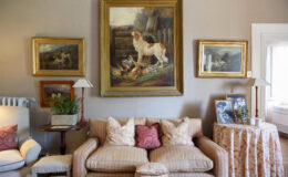 Cozy living room featuring a vintage sofa with decorative pillows, surrounded by framed animal paintings on the walls. A side table with a lamp, potted plant, and photo frames adds to the warm ambiance. Natural light streams in from a nearby window, enhancing the inviting atmosphere.