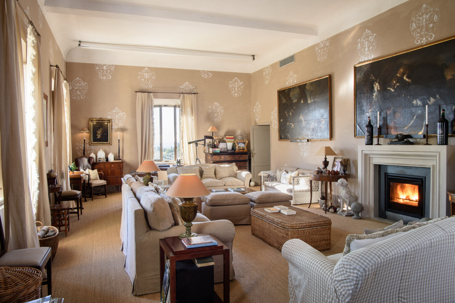 Spacious living room featuring elegant furnishings, a cozy fireplace, and large windows allowing natural light. The decor includes artistic wall paintings and a mix of traditional and modern elements, creating a warm and inviting atmosphere. | Only on SopranoVillas