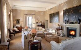 Spacious living room featuring elegant furnishings, a cozy fireplace, and large windows allowing natural light. The decor includes artistic wall paintings and a mix of traditional and modern elements, creating a warm and inviting atmosphere.