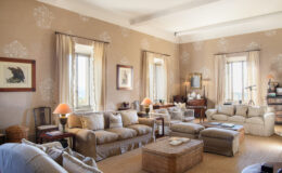 Spacious and elegantly designed living room featuring beige walls, large windows with soft drapes, comfortable seating, a wooden coffee table, and decorative accents, creating a warm and inviting atmosphere.