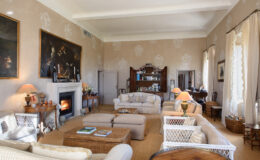 Elegant living room featuring plush sofas, a cozy fireplace, and vintage decor. The room is adorned with artwork and has large windows allowing natural light, creating a warm and inviting atmosphere.