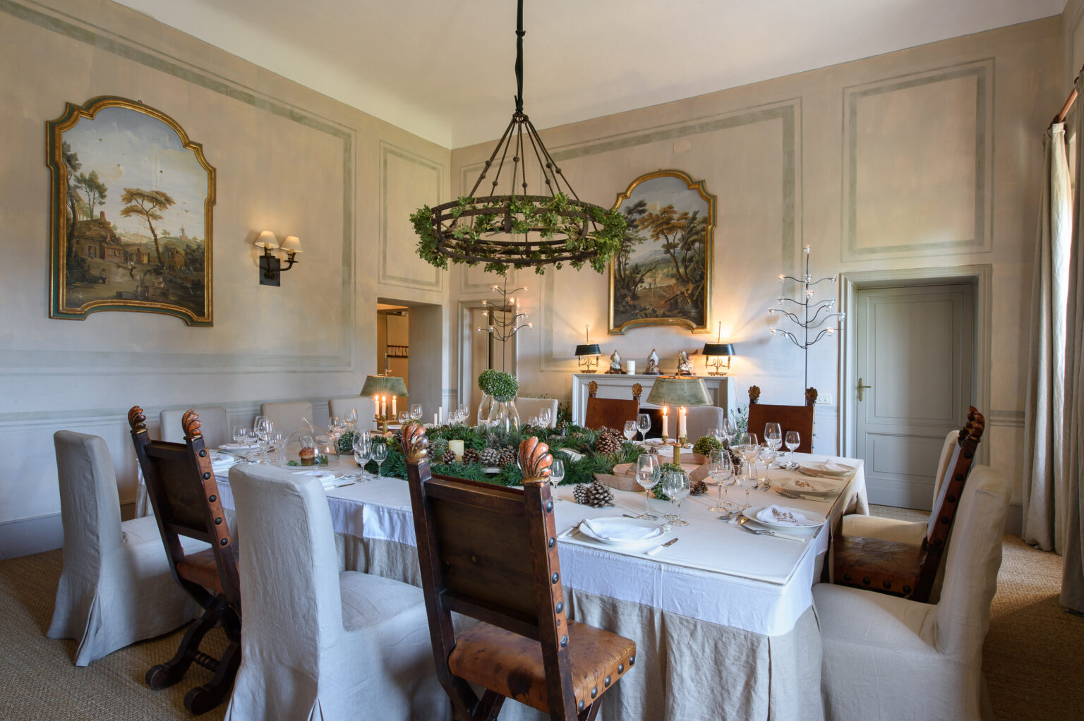 Elegant dining room featuring a large table set for a formal meal, adorned with greenery and decorative tableware. The room showcases classic wall art, a chandelier, and stylish seating, creating a sophisticated atmosphere for gatherings. | Only on SopranoVillas