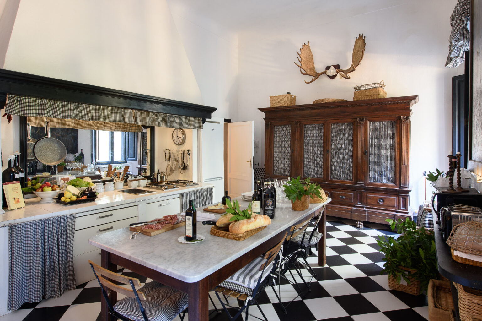 Spacious kitchen with a large marble countertop, black and white checkered flooring, rustic wooden furniture, and decorative elements including a vintage cabinet and wall-mounted antlers. Fresh produce and wine bottles are arranged on the table, enhancing the inviting atmosphere. | Only on SopranoVillas