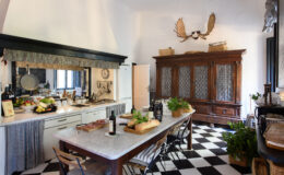 Spacious kitchen with a large marble countertop, black and white checkered flooring, rustic wooden furniture, and decorative elements including a vintage cabinet and wall-mounted antlers. Fresh produce and wine bottles are arranged on the table, enhancing the inviting atmosphere.