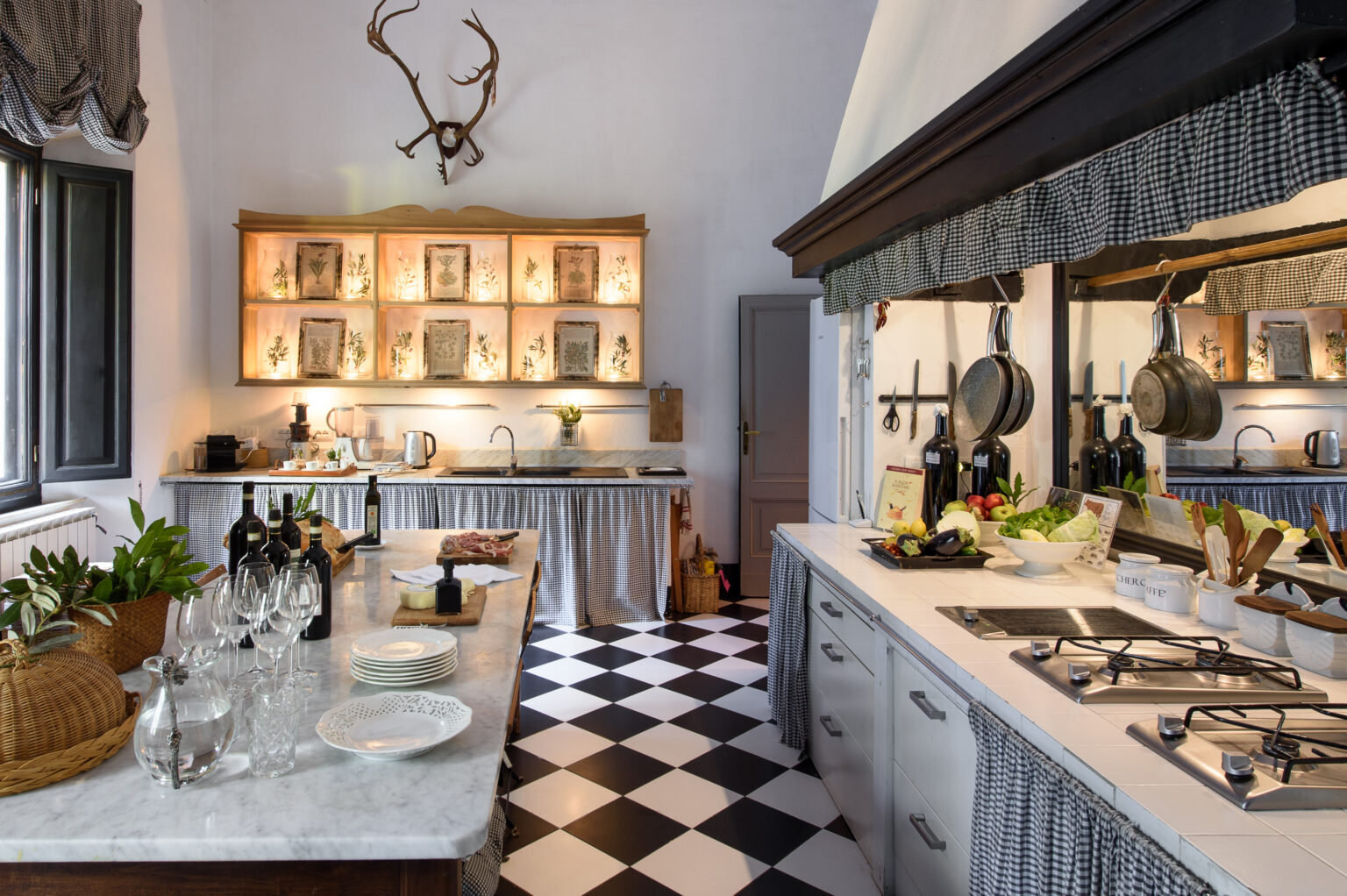 Modern kitchen interior featuring a black and white checkered floor, marble countertops, and wooden cabinetry. Shelves with decorative items and plants add a stylish touch, while stainless steel appliances and cooking utensils emphasize functionality. Natural light enhances the inviting atmosphere. | Only on SopranoVillas