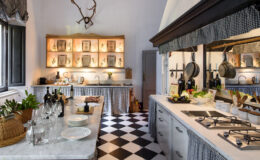 Modern kitchen interior featuring a black and white checkered floor, marble countertops, and wooden cabinetry. Shelves with decorative items and plants add a stylish touch, while stainless steel appliances and cooking utensils emphasize functionality. Natural light enhances the inviting atmosphere.