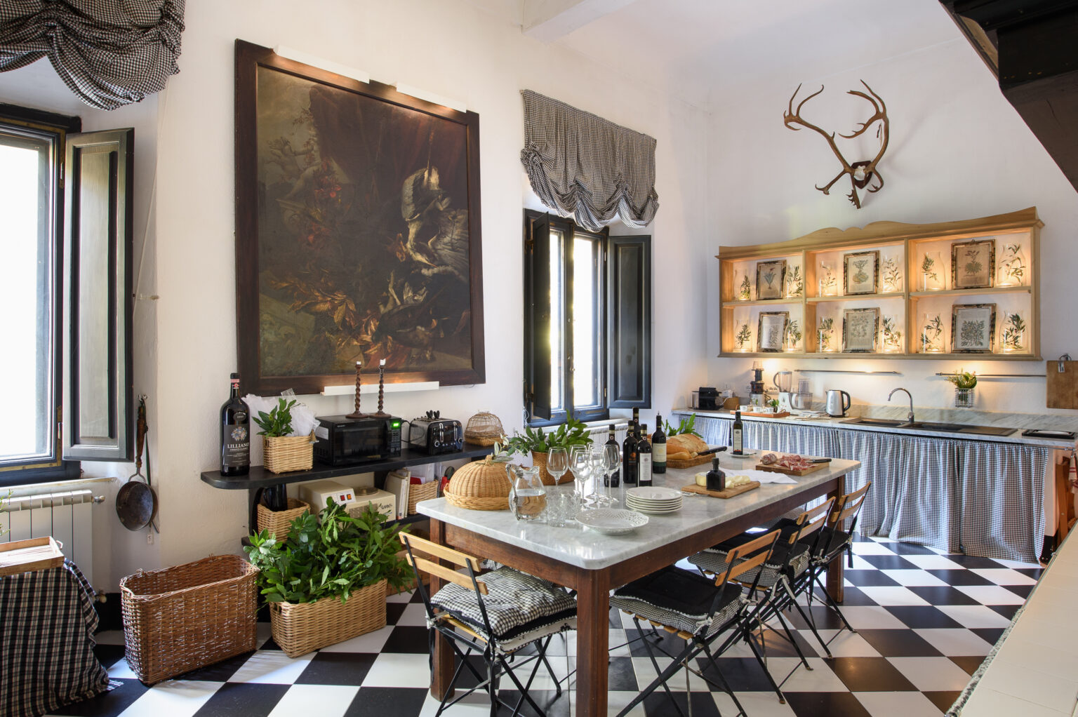 Charming kitchen interior featuring a large painting, wooden shelves with decorative items, a dining table set for a meal, and stylish black and white checkered flooring, complemented by greenery and rustic decor. | Only on SopranoVillas