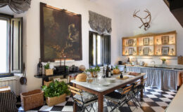 Charming kitchen interior featuring a large painting, wooden shelves with decorative items, a dining table set for a meal, and stylish black and white checkered flooring, complemented by greenery and rustic decor.