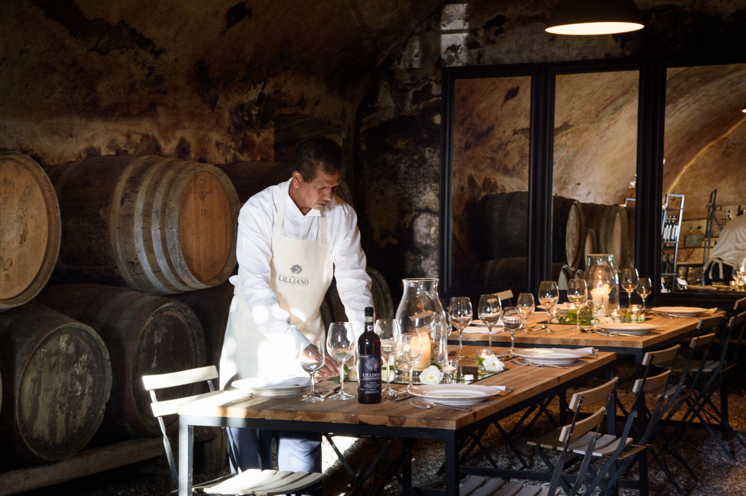 Chef preparing a rustic dining table in a wine cellar, surrounded by wooden barrels and elegant glassware. | Only on SopranoVillas