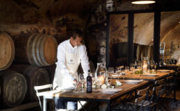 Chef preparing a rustic dining table in a wine cellar, surrounded by wooden barrels and elegant glassware.