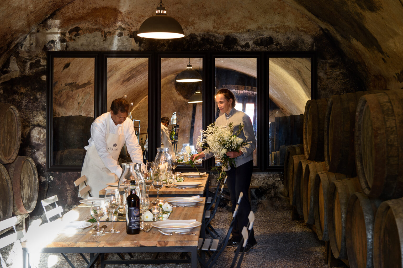Two men preparing a rustic dining table in a wine cellar, surrounded by wine barrels, with elegant table settings and floral arrangements. | Only on SopranoVillas