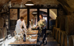 Two men preparing a rustic dining table in a wine cellar, surrounded by wine barrels, with elegant table settings and floral arrangements.