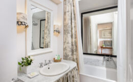 Elegant bathroom featuring a marble countertop sink, decorative wall mirror, and a bathtub with a glass enclosure. Soft, neutral tones and floral curtains enhance the inviting atmosphere.