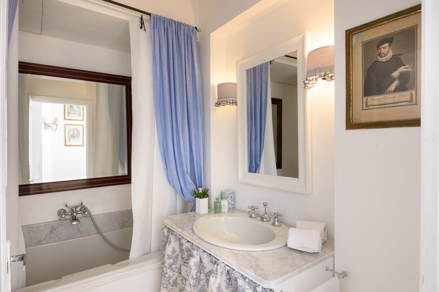 Modern bathroom interior featuring a marble countertop, a bathtub with a shower curtain, a large mirror, and decorative wall sconces. The space is accented with light blue curtains and framed art, creating a serene atmosphere. | Only on SopranoVillas