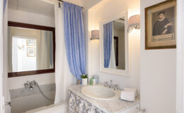 Modern bathroom interior featuring a marble countertop, a bathtub with a shower curtain, a large mirror, and decorative wall sconces. The space is accented with light blue curtains and framed art, creating a serene atmosphere.