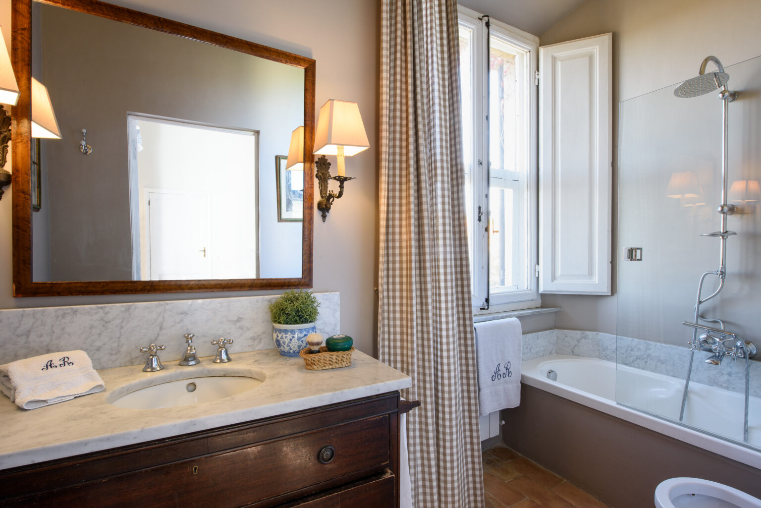 Modern bathroom with a marble vanity, decorative wall mirror, and a bathtub with a glass shower enclosure, featuring natural light from a window and elegant wall sconces. | Only on SopranoVillas