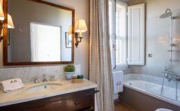 Modern bathroom with a marble vanity, decorative wall mirror, and a bathtub with a glass shower enclosure, featuring natural light from a window and elegant wall sconces.