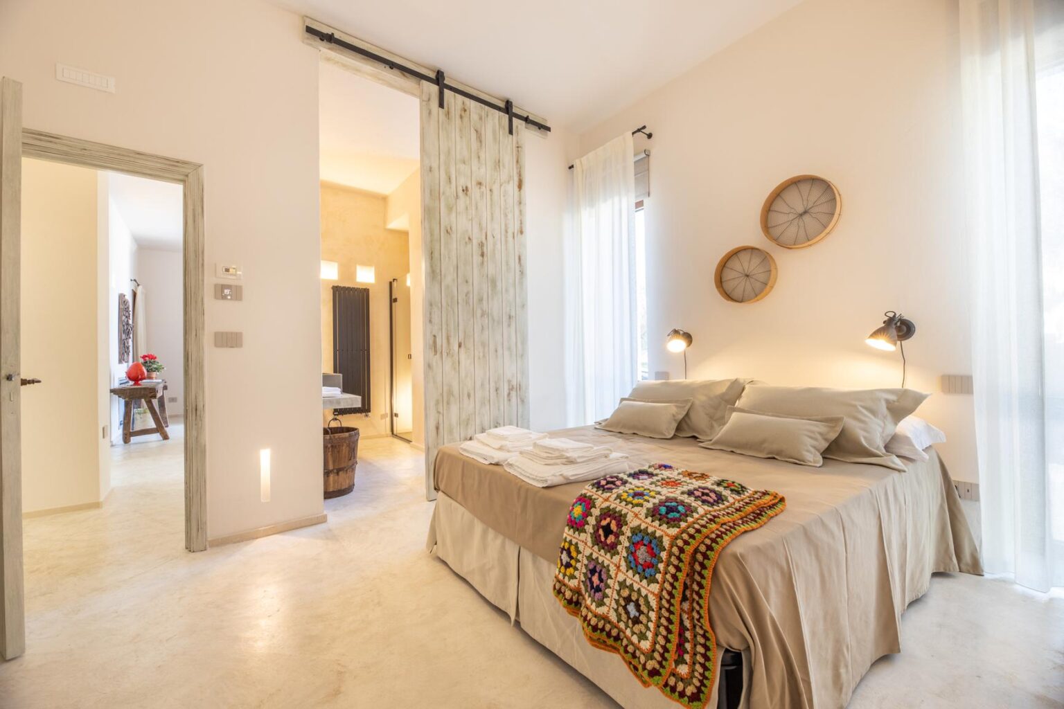 Cozy bedroom featuring a neatly made bed with decorative pillows and a colorful crocheted blanket, illuminated by stylish wall lamps. The room has large windows with sheer curtains, and a sliding barn door leads to a modern bathroom area, enhancing the open and airy feel of the space. | Only on SopranoVillas