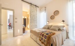 Cozy bedroom featuring a neatly made bed with decorative pillows and a colorful crocheted blanket, illuminated by stylish wall lamps. The room has large windows with sheer curtains, and a sliding barn door leads to a modern bathroom area, enhancing the open and airy feel of the space.