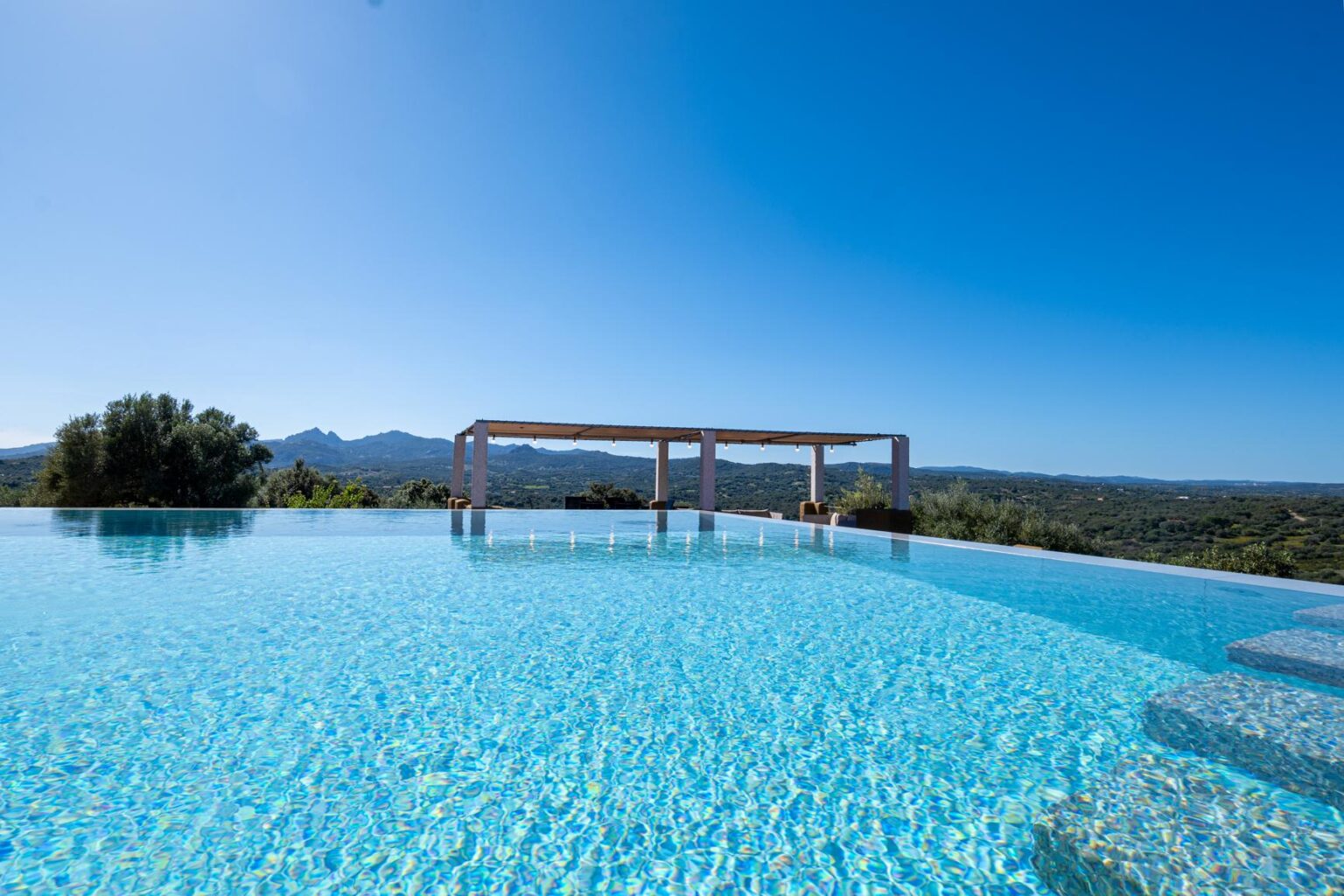 Stunning infinity pool overlooking a mountainous landscape under a clear blue sky, featuring modern architecture and tranquil water. | Only on SopranoVillas