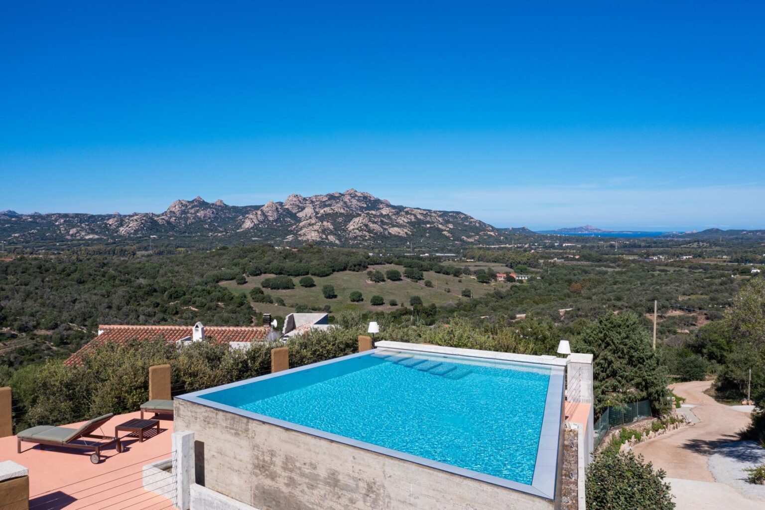 Stunning view of an infinity pool overlooking lush green hills and rocky mountains under a clear blue sky, showcasing a serene outdoor space ideal for relaxation. | Only on SopranoVillas