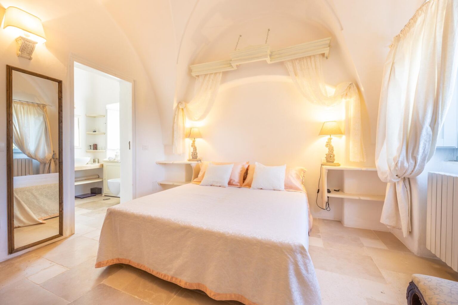 Bright and airy bedroom featuring a double bed with soft linens, elegant lamps, and a large mirror. The room has light-colored walls and a cozy atmosphere, with a doorway leading to a modern bathroom. Ideal for relaxation and comfort. | Only on SopranoVillas