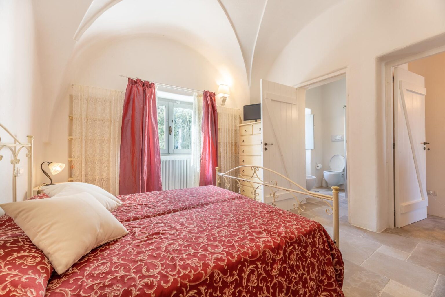 Cozy bedroom featuring two twin beds with red patterned bedding, soft pillows, and elegant drapes. The room has arched ceilings, natural light from a window, and a minimalist decor style, creating a warm and inviting atmosphere. | Only on SopranoVillas