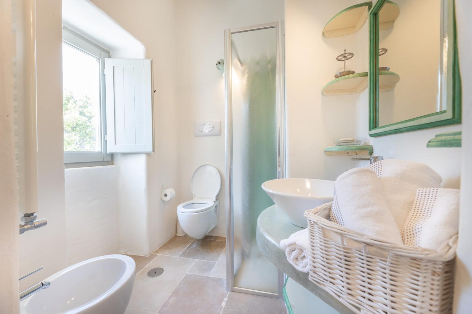Bright and modern bathroom featuring a glass shower, white toilet, stylish sink, and a basket of towels, with natural light streaming in through a window. | Only on SopranoVillas