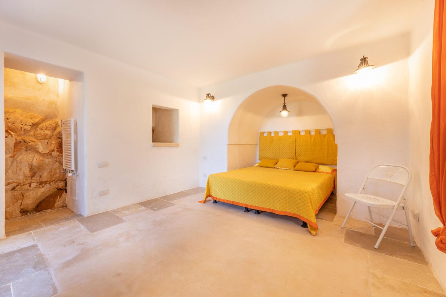 Bright and airy bedroom featuring a yellow bedspread, minimalist decor, and warm lighting, with a stone wall accent and a white folding chair. Ideal for a cozy retreat. | Only on SopranoVillas