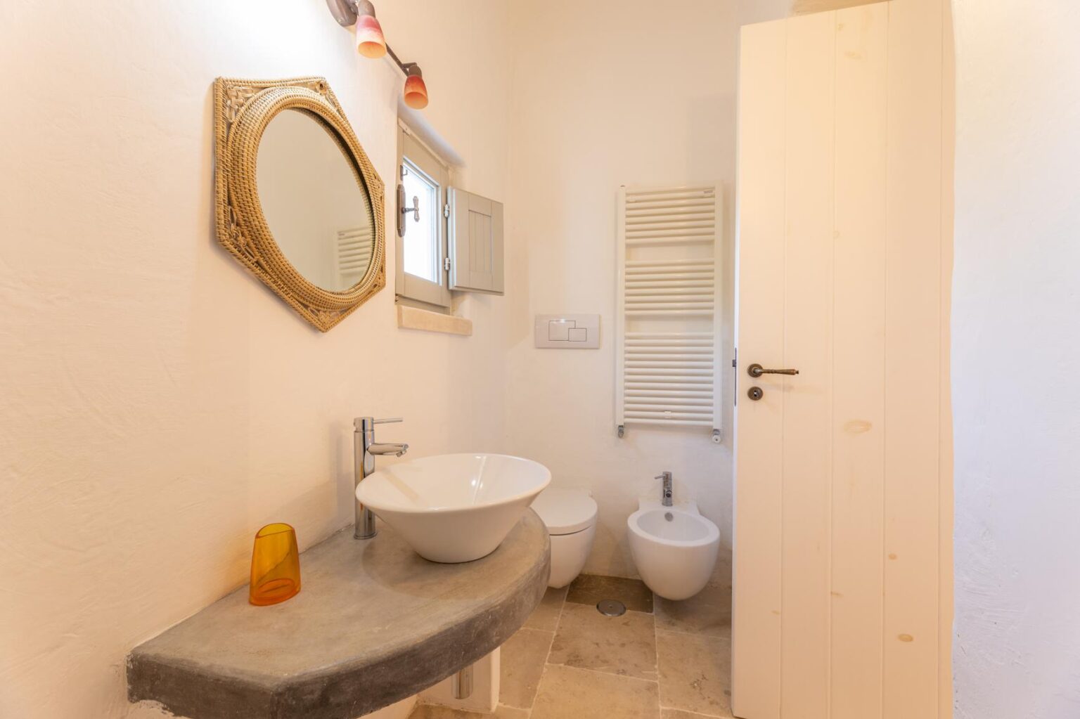 Modern bathroom interior featuring a round sink, wall-mounted mirror, bidet, and towel radiator against a light-colored wall. The design emphasizes cleanliness and simplicity with natural light enhancing the space. | Only on SopranoVillas