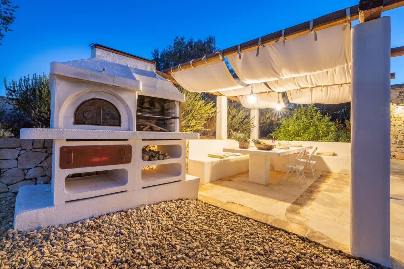 Outdoor kitchen featuring a traditional stone pizza oven, dining area, and shaded seating under a pergola, surrounded by lush greenery at dusk. | Only on SopranoVillas