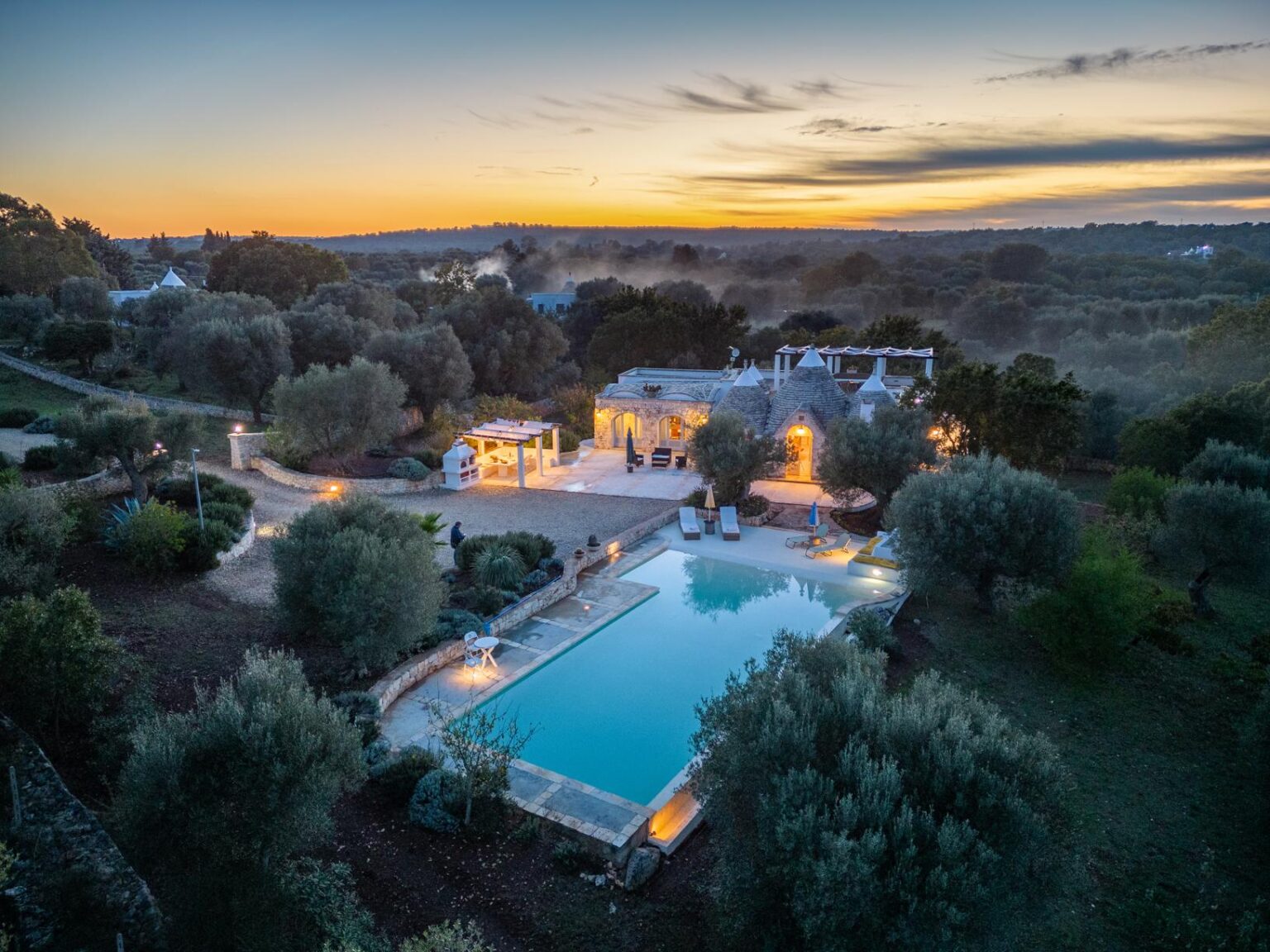 Aerial view of a luxurious villa with a swimming pool surrounded by olive trees at sunset, highlighting the tranquil landscape and charming architecture. | Only on SopranoVillas