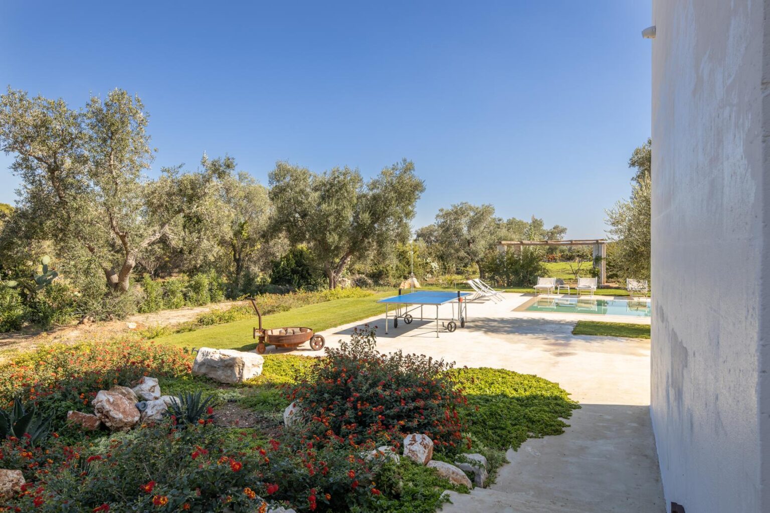 Scenic view of a landscaped garden featuring olive trees, colorful flowers, and a swimming pool, under a clear blue sky. | Only on SopranoVillas