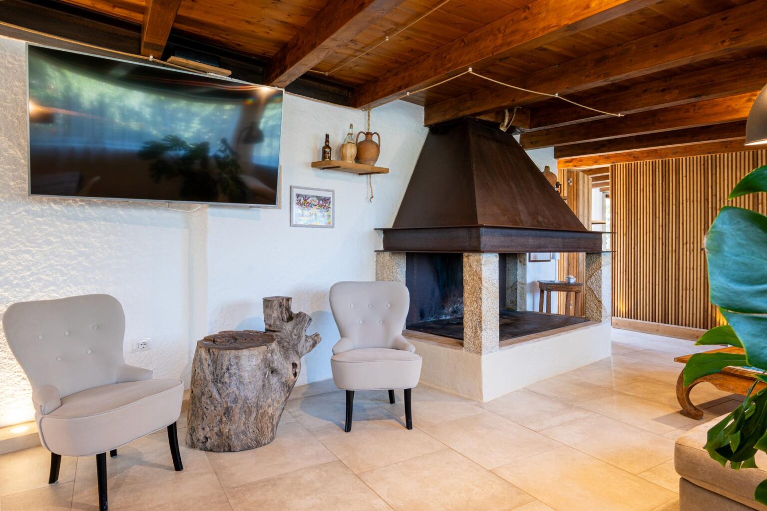 Cozy living room featuring a modern fireplace, two elegant upholstered chairs, a rustic wooden side table, and a flat-screen TV mounted on the wall, set against a backdrop of wooden beams and textured walls. Perfect for a warm and inviting atmosphere. | Only on SopranoVillas
