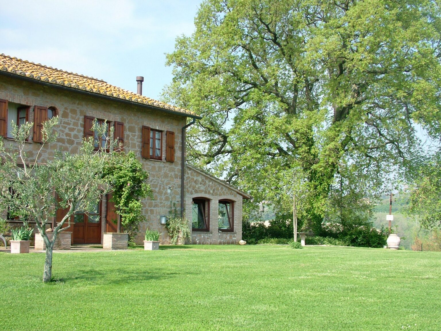 Charming stone farmhouse with red shutters surrounded by lush green lawn and trees, set against a clear blue sky. | Only on SopranoVillas