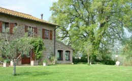 Charming stone farmhouse with red shutters surrounded by lush green lawn and trees, set against a clear blue sky.