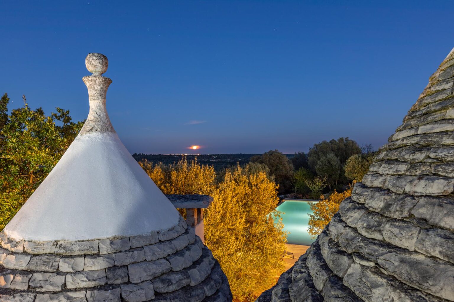 Trulli rooftops against a twilight sky, showcasing the warm colors of autumn foliage and a distant sunset in the background. | Only on SopranoVillas