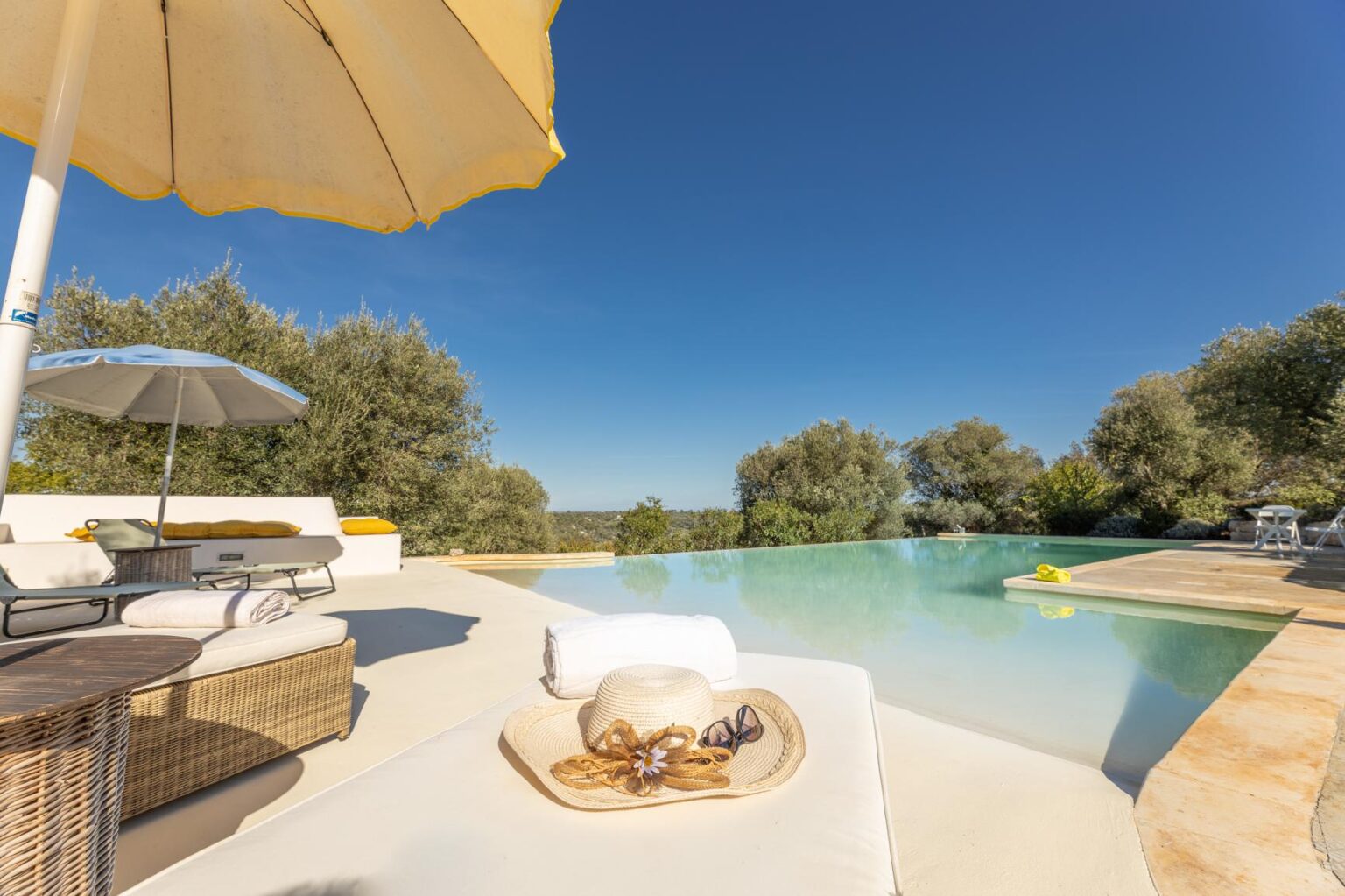 Luxury outdoor pool area with a sun umbrella, comfortable lounge chairs, and a serene landscape of olive trees under a clear blue sky. | Only on SopranoVillas