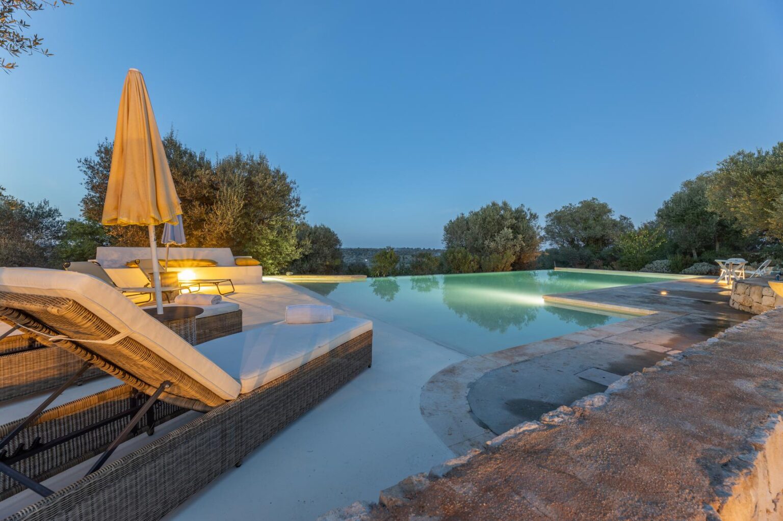 Luxury infinity pool with lounge chairs and an umbrella, surrounded by lush greenery at dusk, offering a serene outdoor retreat. | Only on SopranoVillas