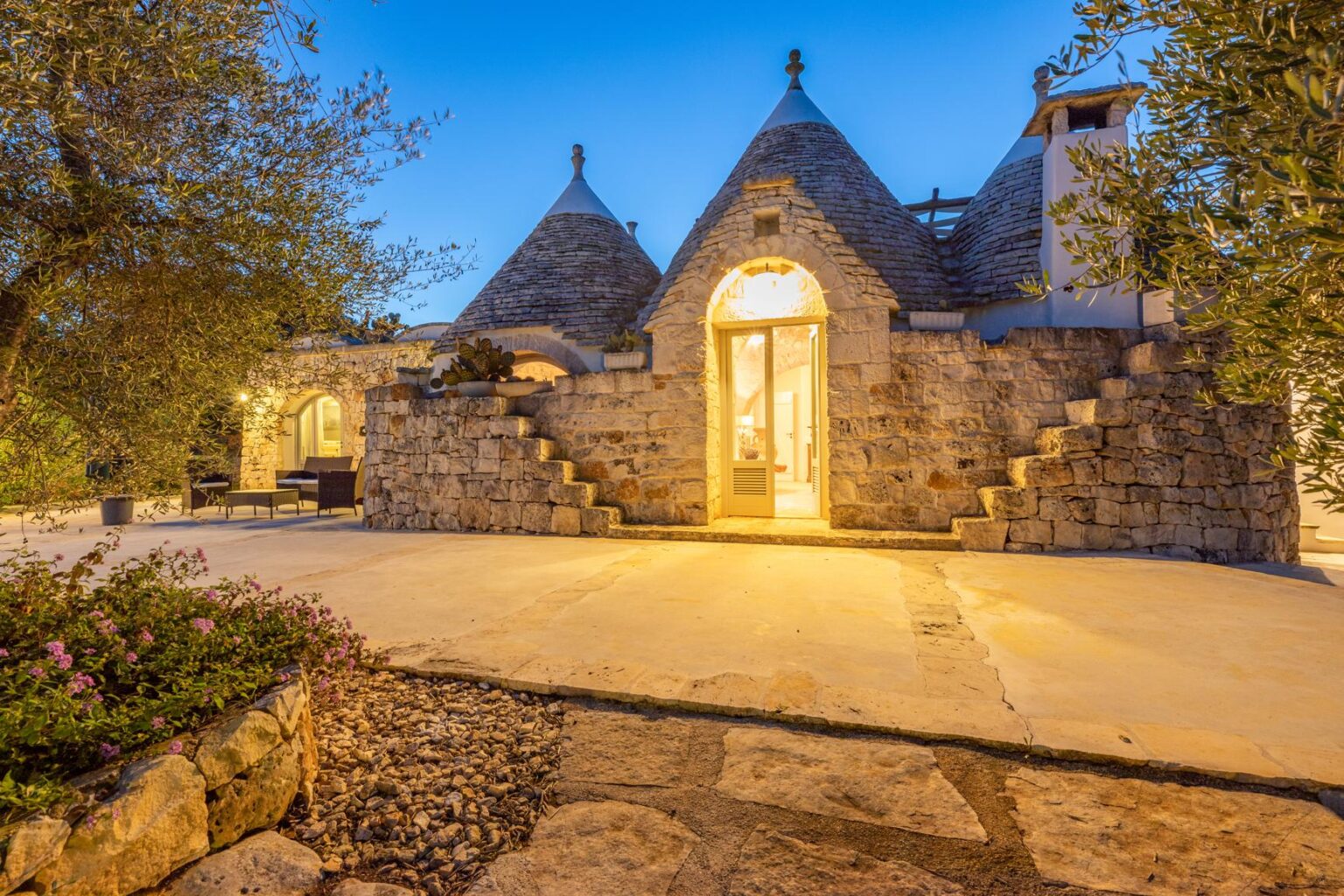Trullo-style house exterior illuminated at dusk, featuring traditional conical roofs and stone walls, surrounded by lush greenery and flowering plants. | Only on SopranoVillas