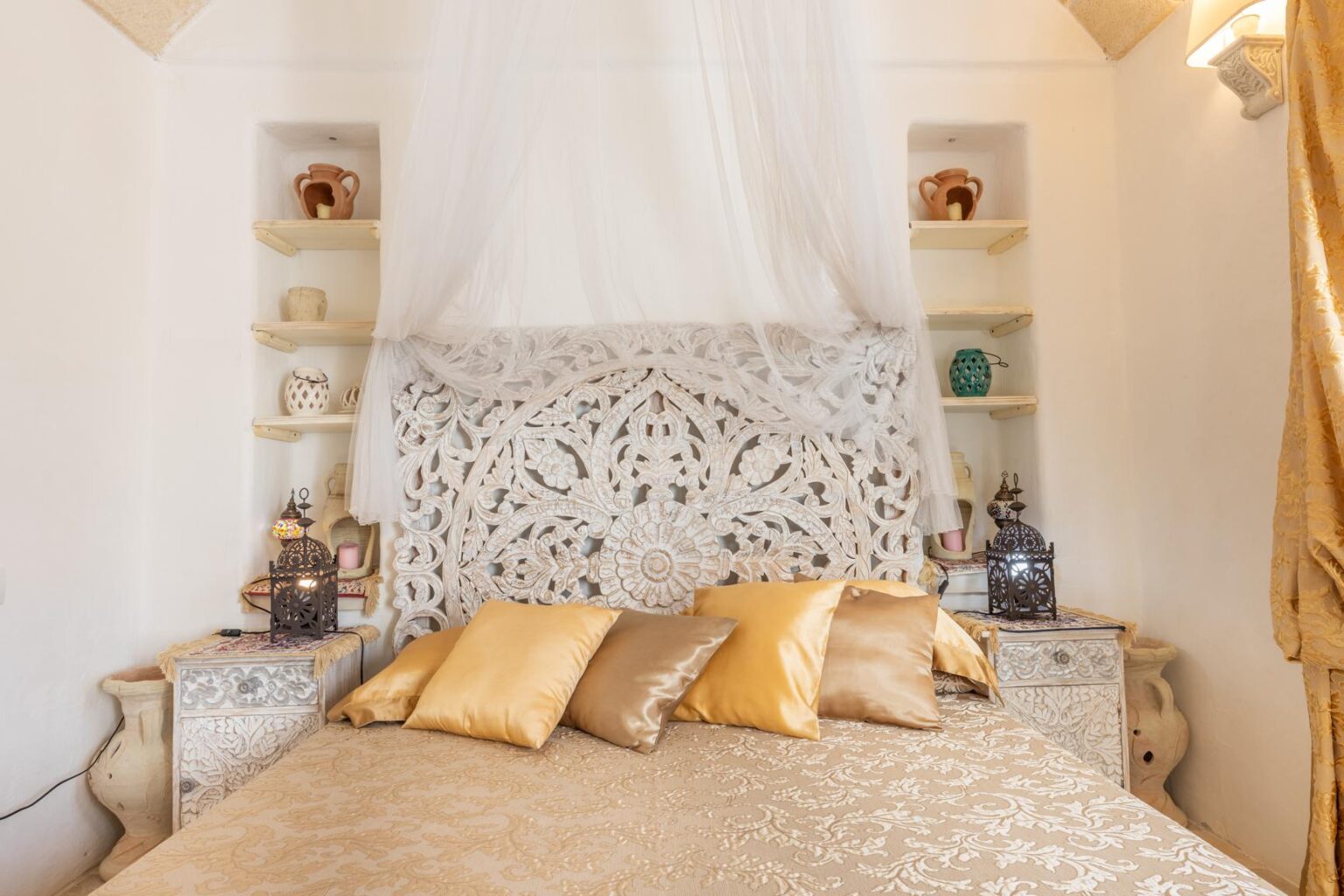 Elegant bedroom featuring a intricately carved wooden headboard draped with a sheer canopy, complemented by luxurious gold and beige decorative pillows. Shelves display various decorative pottery and lanterns, creating a cozy and inviting atmosphere. | Only on SopranoVillas