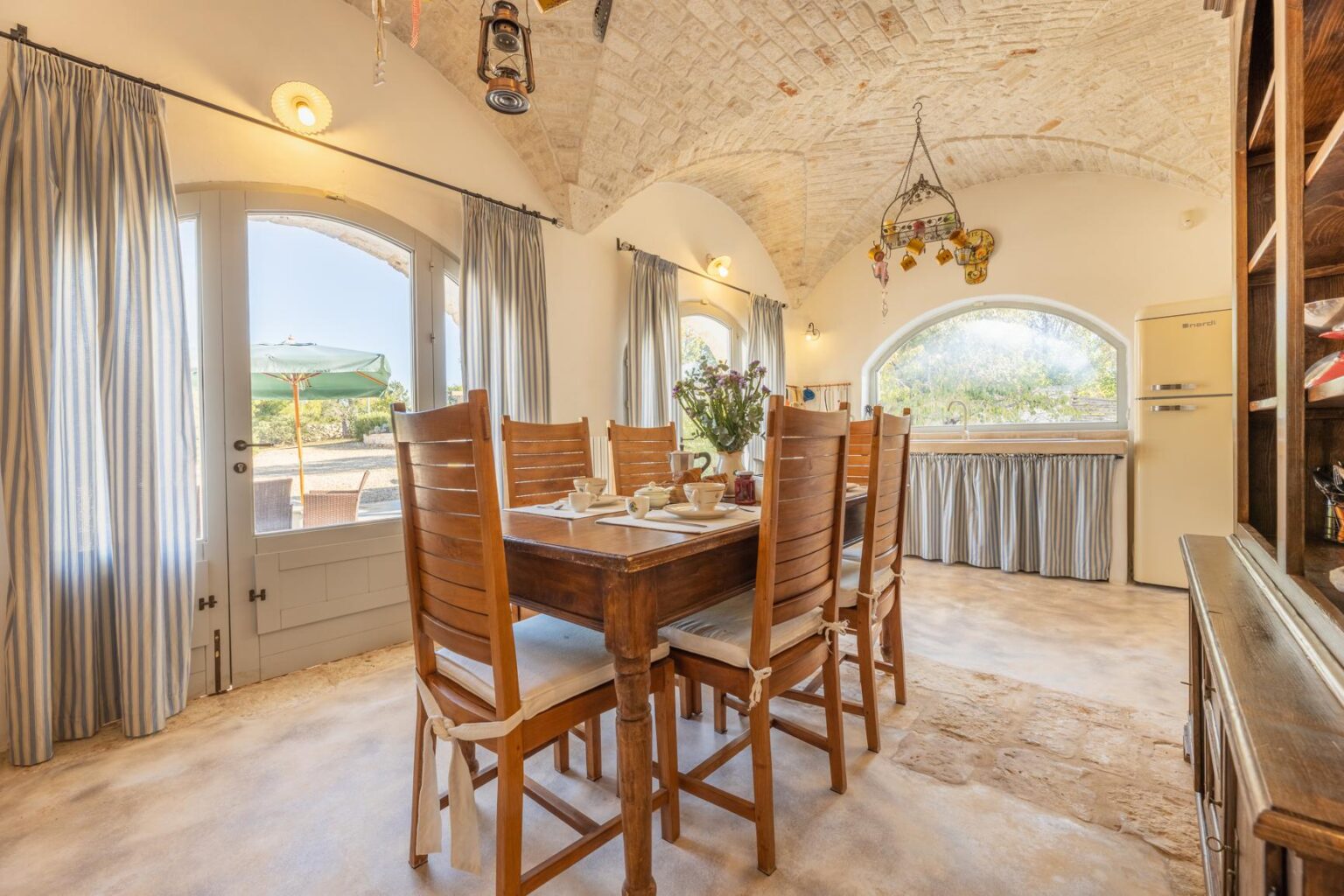 Bright and airy dining area featuring a wooden table set for breakfast, with six chairs, a vase of flowers, and a view of the outdoors through large windows. The room has a rustic charm with a vaulted ceiling, light-colored walls, and a vintage refrigerator. Natural light pours in, enhancing the inviting atmosphere. | Only on SopranoVillas