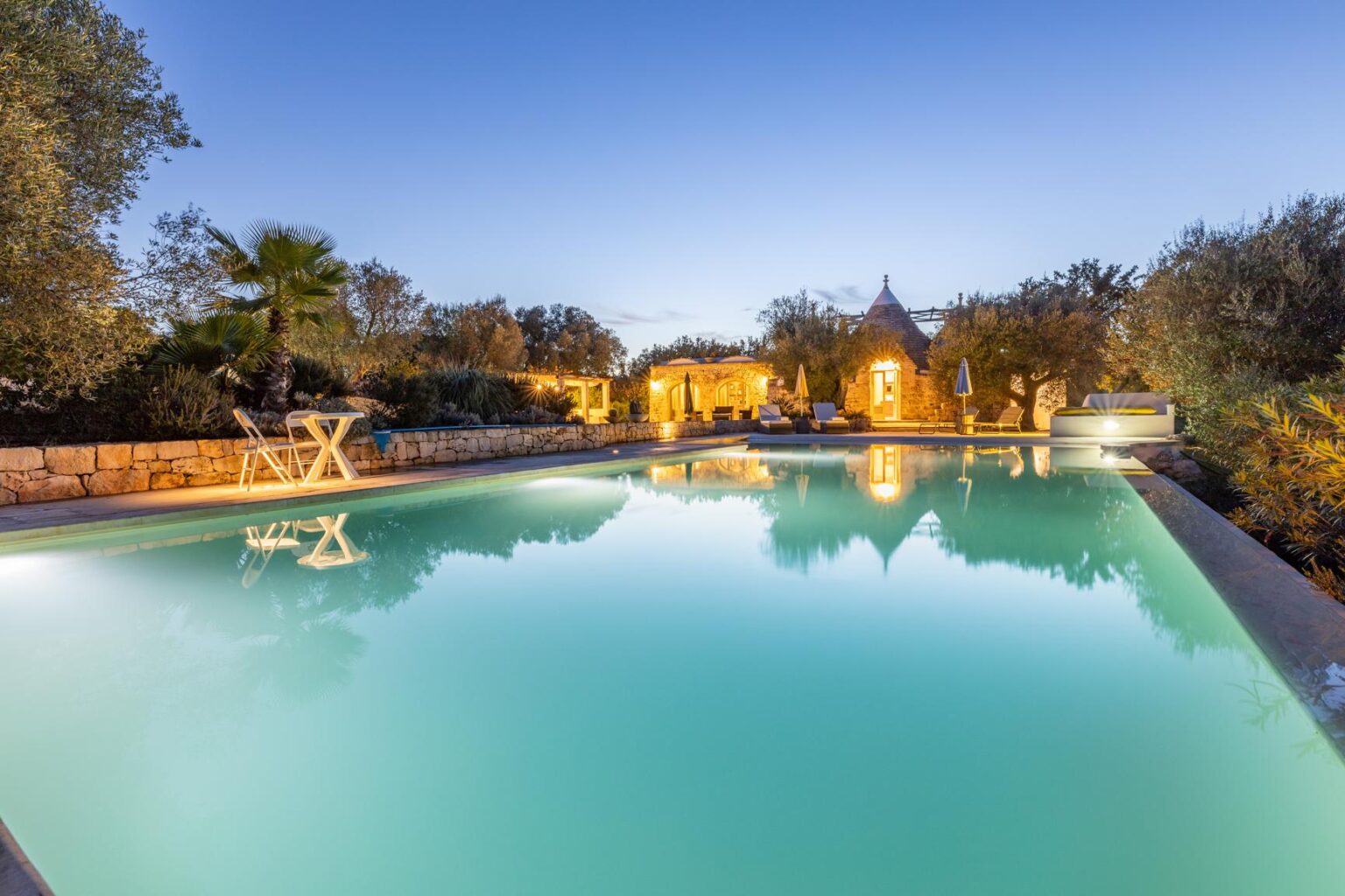 Serene evening view of a luxury pool surrounded by lush greenery and a stone patio, featuring comfortable seating and a charming stone building, perfect for relaxation and outdoor gatherings. | Only on SopranoVillas