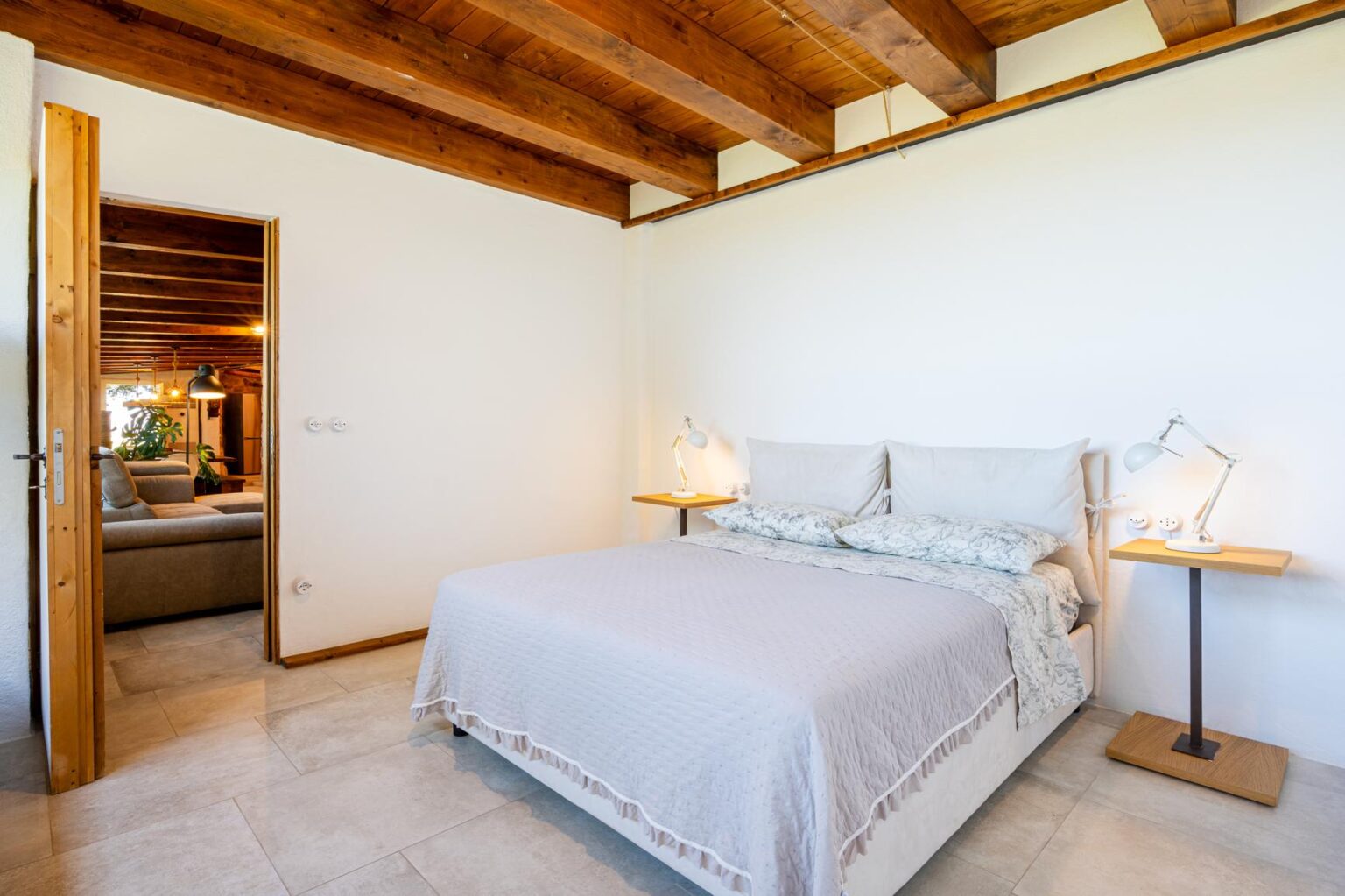 Cozy bedroom featuring a queen-sized bed with light bedding, two bedside lamps, and modern decor. The room has wooden beams on the ceiling and a doorway leading to a living area with a sofa and plants, creating an inviting atmosphere. | Only on SopranoVillas