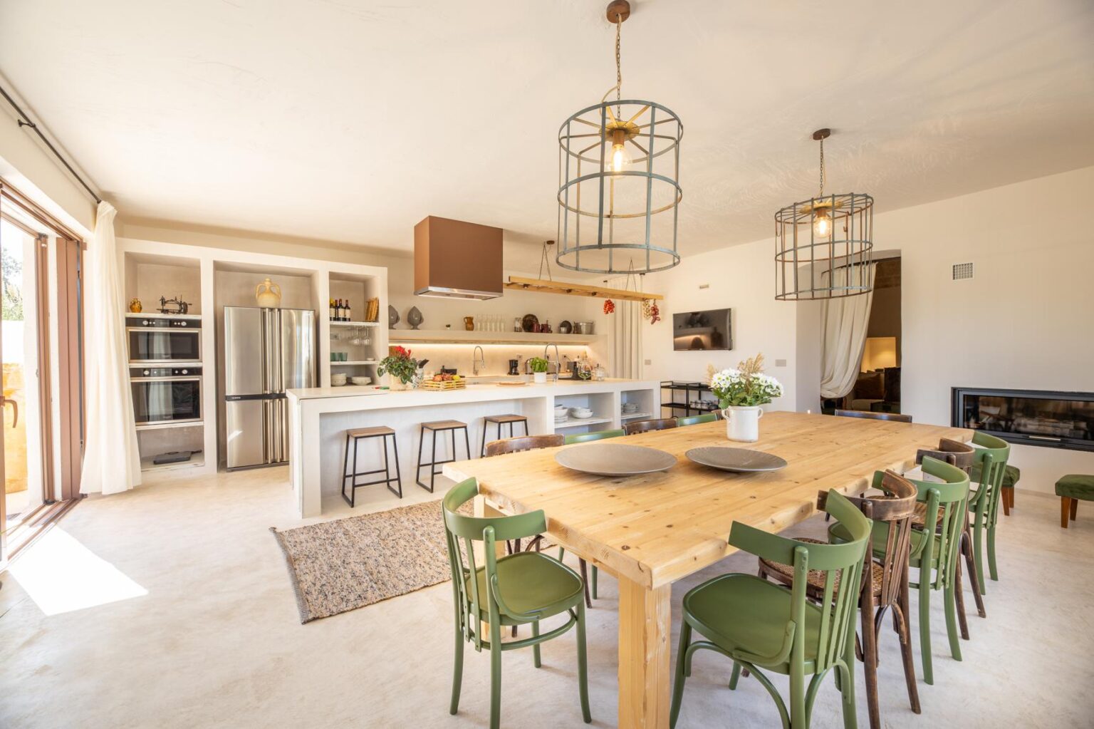 Bright and airy modern kitchen with a spacious dining area featuring a large wooden table surrounded by green chairs, pendant lighting, and open shelving, creating a welcoming atmosphere for family gatherings. | Only on SopranoVillas