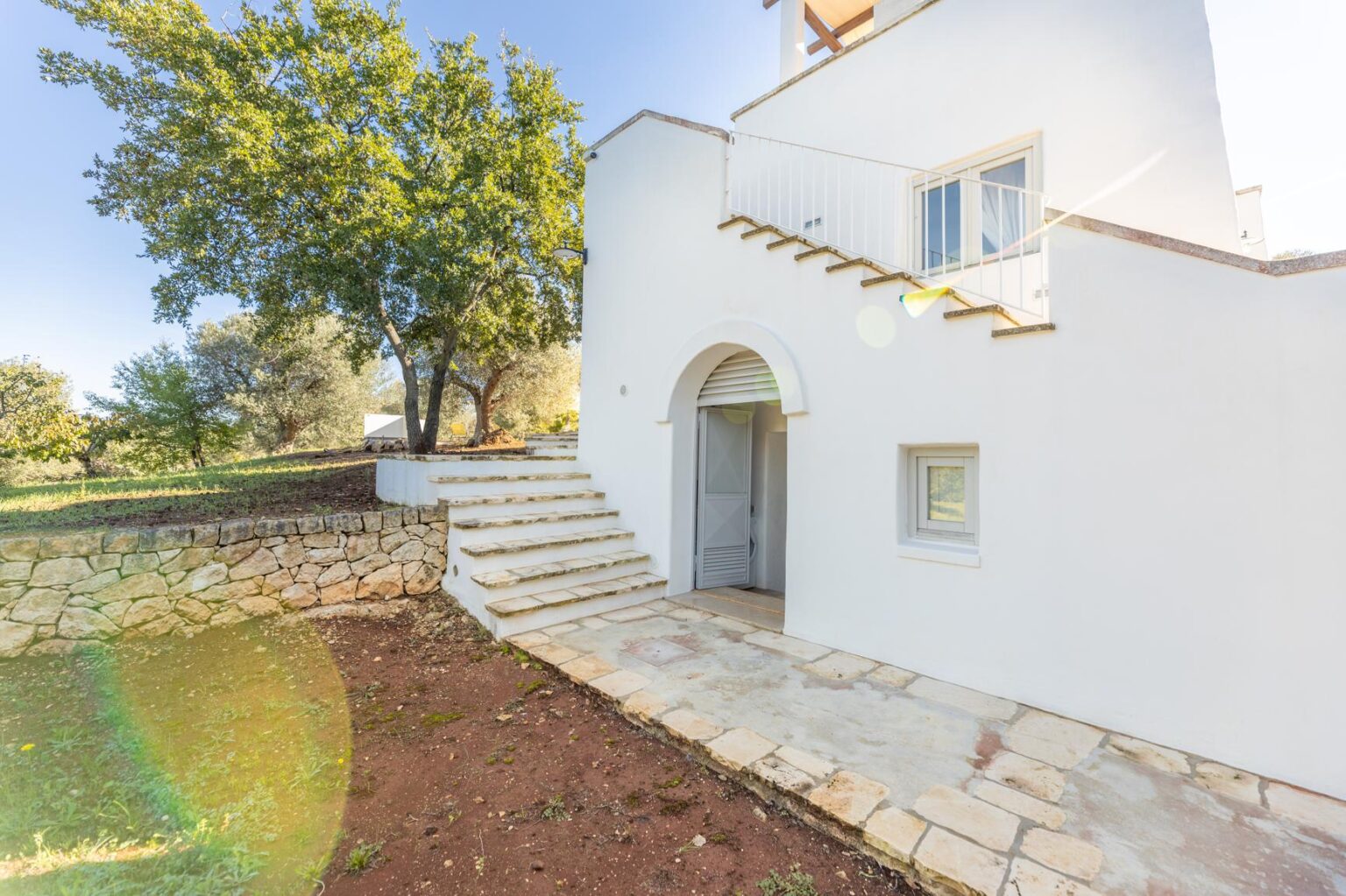 Modern white house with stone steps leading to an arched doorway, surrounded by greenery and sunlight. Ideal for showcasing outdoor living and architectural design. | Only on SopranoVillas