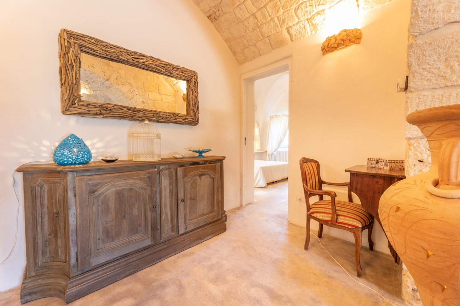 Elegant interior hallway featuring a wooden sideboard with decorative items, a vintage chair, and a stone wall. The warm lighting enhances the inviting atmosphere, showcasing a blend of rustic and modern design elements. | Only on SopranoVillas